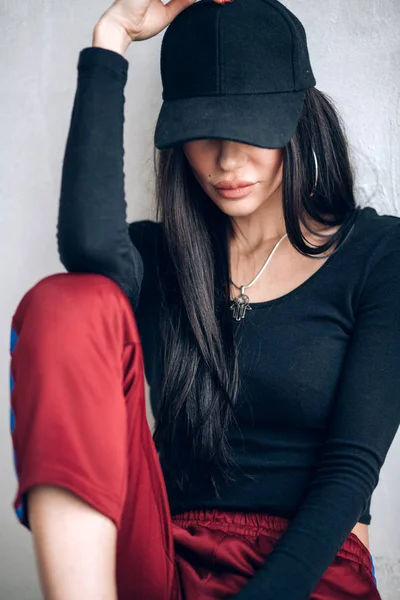 Portrait of a brunette girl in a cap and sportswear. Natural light. Red sweatpants, black top, black cap.