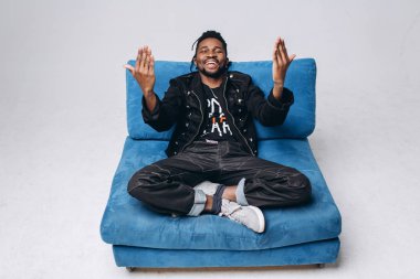 Bir çekici gülümseyen siyah adam bir siyah ceket ve gri arka plan mavi kanepede sidin üzerinde siyah pantolon