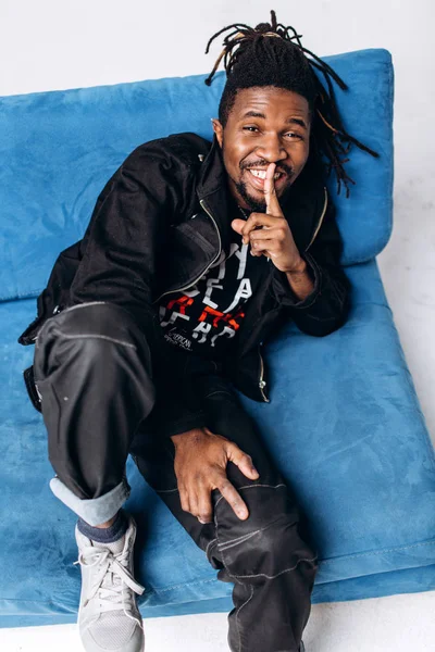 One attractive smiling black man in a black jacket and black pants on a gray background sidin on a blue sofa