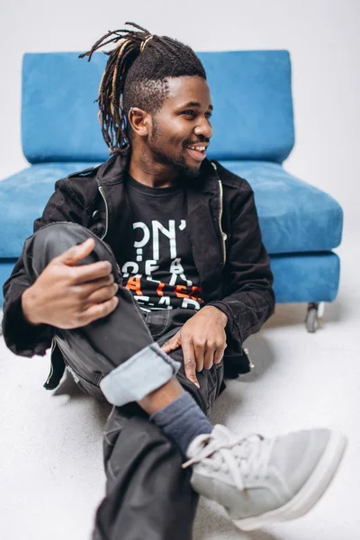 One attractive smiling black man in a black jacket and black pants on a gray background sidin on a blue sofa
