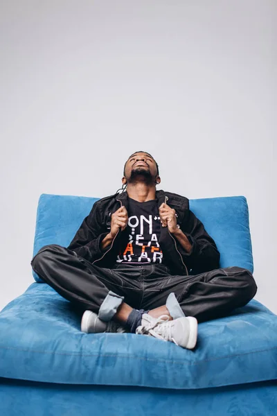 One attractive smiling black man in a black jacket and black pants on a gray background sidin on a blue sofa