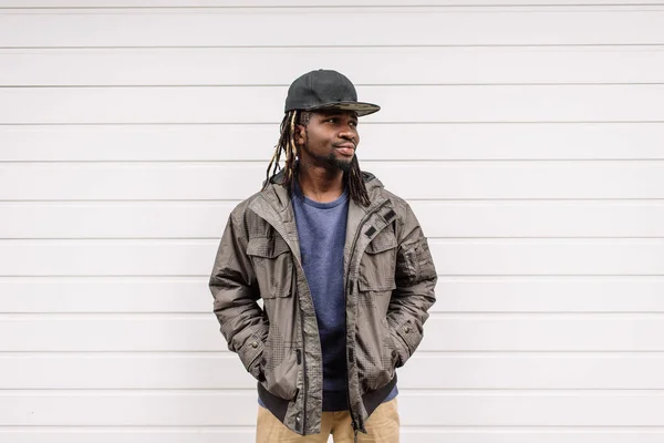 Pěkný africký Američan s dredy v hnědých kalhotách a modrý svetr v černé čepici na hlavě na pozadí domů, město — Stock fotografie
