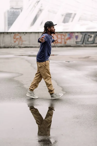 Bonito afro-americano com dreadlocks em calças marrons e uma camisola azul em um boné preto em sua cabeça contra o fundo das casas, cidade — Fotografia de Stock