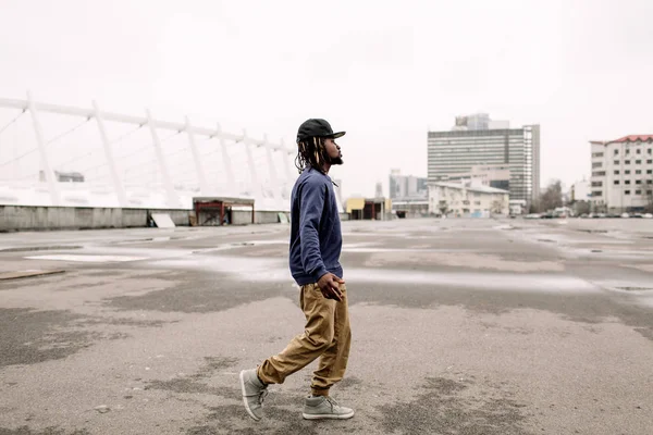 Bonito afro-americano com dreadlocks em calças marrons e uma camisola azul em um boné preto em sua cabeça contra o fundo das casas, cidade — Fotografia de Stock