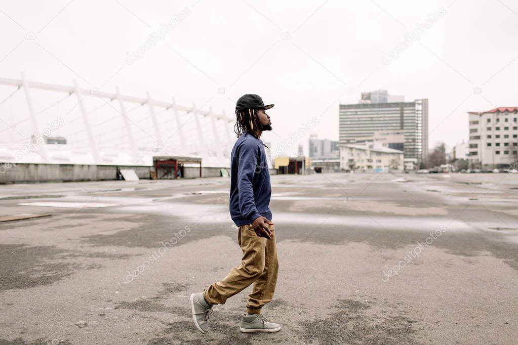 Handsome African American with dreadlocks in brown trousers and a blue sweater in a black cap on his head against the background of houses, city