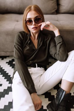Güzel tatlı kız kanepede yatıyor. Kısa kırmızı saçlı Altın şık model, dolgun dudaklar, güneş gözlüğü, rahat giyim, kostüm, kıyafet, tulum. Model testi, reklam gözlükleri, giysiler