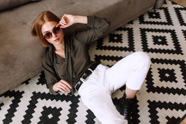 Güzel tatlı kız kanepede yatıyor. Kısa kırmızı saçlı Altın şık model, dolgun dudaklar, güneş gözlüğü, rahat giyim, kostüm, kıyafet, tulum. Model testi, reklam gözlükleri, giysiler