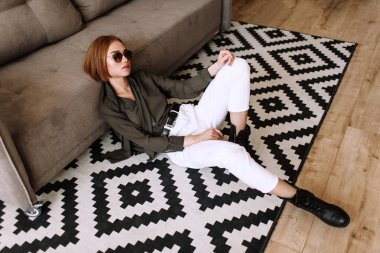 Güzel tatlı kız kanepede yatıyor. Kısa kırmızı saçlı Altın şık model, dolgun dudaklar, güneş gözlüğü, rahat giyim, kostüm, kıyafet, tulum. Model testi, reklam gözlükleri, giysiler