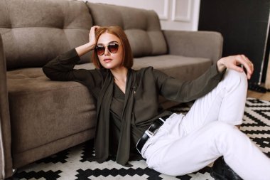 Güzel tatlı kız kanepede yatıyor. Kısa kırmızı saçlı Altın şık model, dolgun dudaklar, güneş gözlüğü, rahat giyim, kostüm, kıyafet, tulum. Model testi, reklam gözlükleri, giysiler