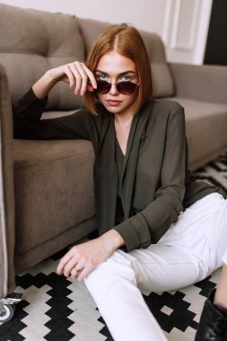 Güzel tatlı kız kanepede yatıyor. Kısa kırmızı saçlı Altın şık model, dolgun dudaklar, güneş gözlüğü, rahat giyim, kostüm, kıyafet, tulum. Model testi, reklam gözlükleri, giysiler