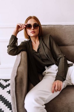 Güzel tatlı kız kanepede yatıyor. Kısa kırmızı saçlı Altın şık model, dolgun dudaklar, güneş gözlüğü, rahat giyim, kostüm, kıyafet, tulum. Model testi, reklam gözlükleri, giysiler