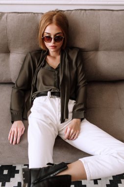 Güzel tatlı kız kanepede yatıyor. Kısa kırmızı saçlı Altın şık model, dolgun dudaklar, güneş gözlüğü, rahat giyim, kostüm, kıyafet, tulum. Model testi, reklam gözlükleri, giysiler