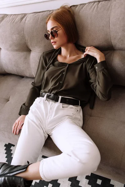 Beautiful sweet girl lying on the sofa. Golden stylish model with short red hair, plump lips, sunglasses, casual clothing, costume, outfit, overalls. Model test, advertising glasses, clothes — Stock Photo, Image