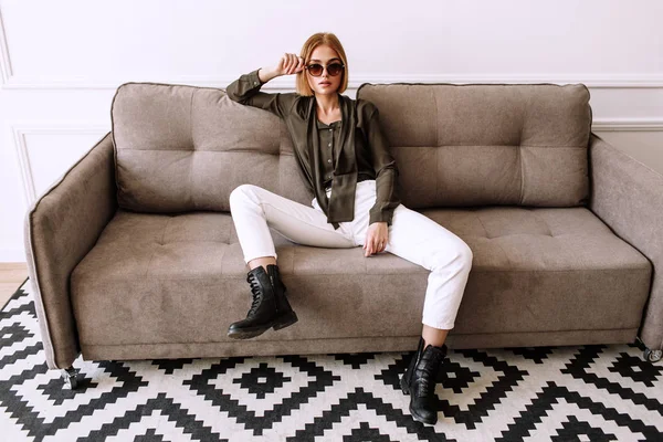 Schöne süße Mädchen auf dem Sofa liegend. goldenes stylisches Model mit kurzen roten Haaren, dicken Lippen, Sonnenbrille, lässiger Kleidung, Kostüm, Outfit, Overalls. Modellversuch, Werbebrille, Kleidung — Stockfoto