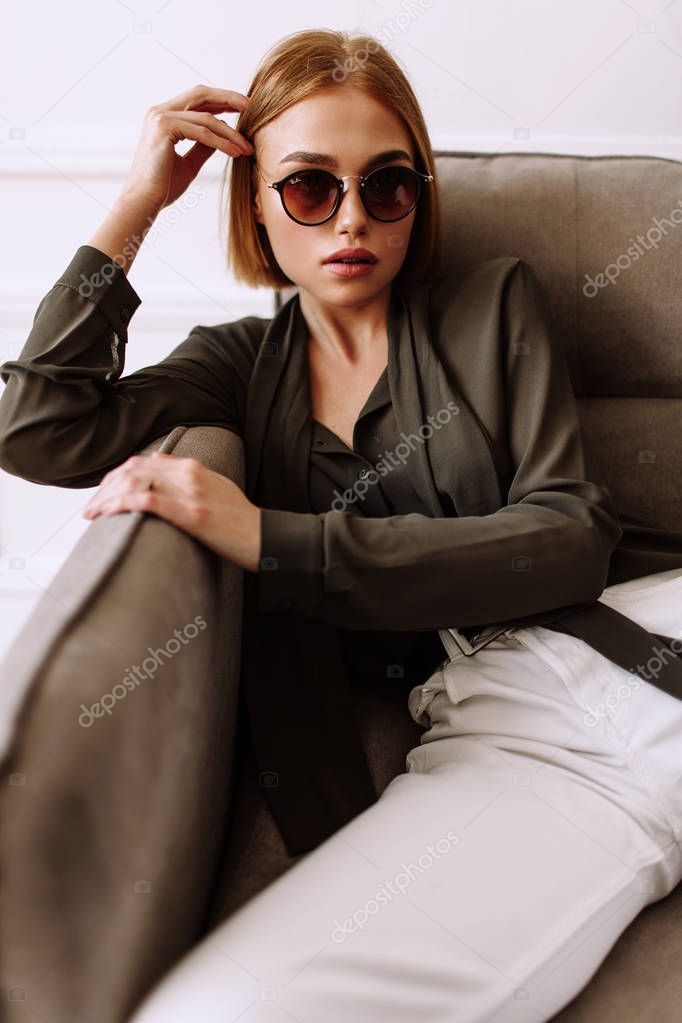 Beautiful sweet girl lying on the sofa. Golden stylish model with short red hair, plump lips, sunglasses, casual clothing, costume, outfit, overalls. Model test, advertising glasses, clothes