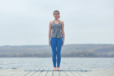 Beautiful young woman practices yoga asana Tadasana - Mountain pose on the wooden deck near the lake. clipart