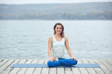Güzel ve genç birbayan uygulamaları yoga asana Padmasana - Lotus poz göl yakınındaki ahşap güverte