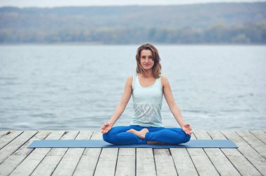 Güzel ve genç birbayan uygulamaları yoga asana Padmasana - Lotus poz göl yakınındaki ahşap güverte