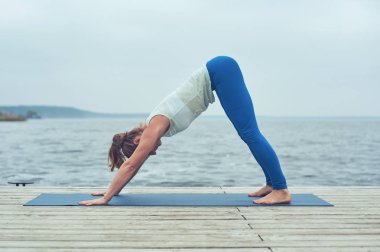 Güzel ve genç birbayan uygulamaları yoga asana göl yakınındaki ahşap güverte'deki aşağı doğru bakan köpek