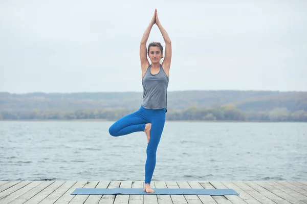 Красивая молодая женщина практикует йогу asana Parshva Vrikshasana - Поза дерева на деревянной палубе возле озера — стоковое фото