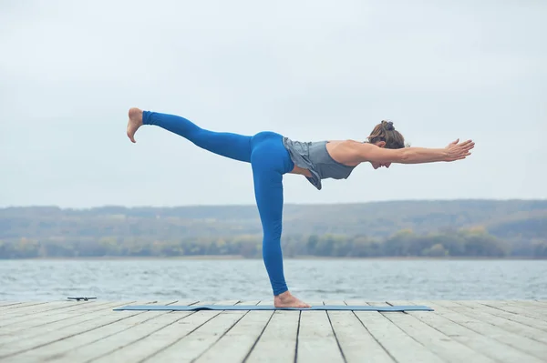 Красивая молодая женщина практикует йогу asana Virabhadrasana 3 - воин позировать 3 на деревянной палубе возле озера — стоковое фото