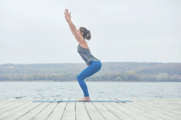 Красивая молодая женщина практикует йогу asana Utkatasana - Поза стула на деревянной палубе возле озера — стоковое фото