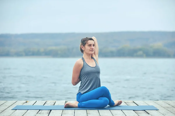 Красивая молодая женщина практикует йогу asana Gomukhasana - Cow face pose on the wooden deck near the lake — стоковое фото