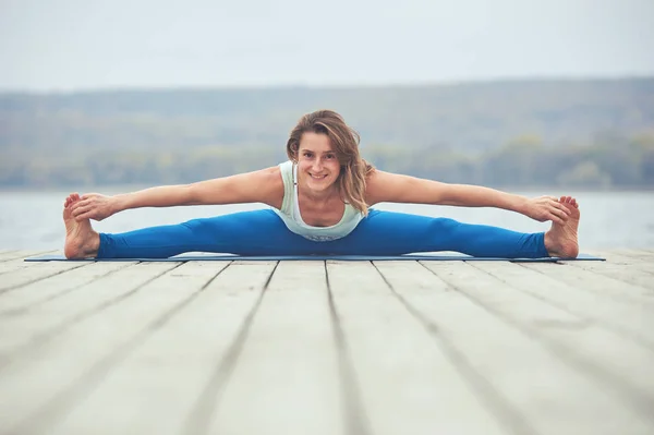 Beautiful young woman practices yoga asana Samakonasana - Straight angle pose on the wooden deck near the lake