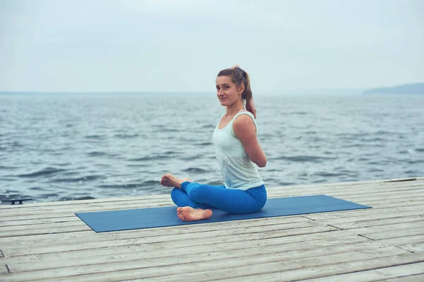 Красивая молодая женщина практикует йогу asana Gomukhasana - Cow face pose on the wooden deck near the lake — стоковое фото