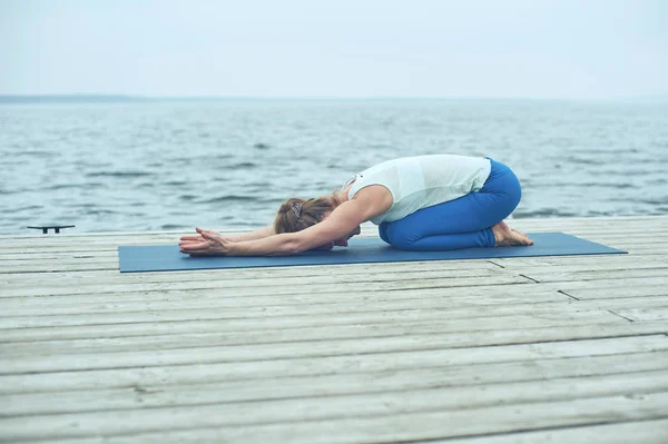 Красивая молодая женщина практикует йогу asana Balasana - дети позируют на деревянной палубе возле озера — стоковое фото