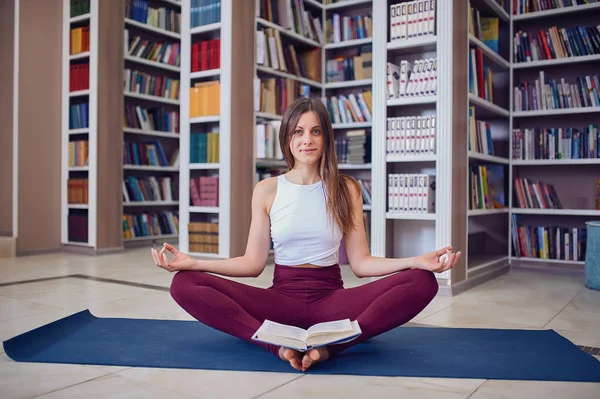 Beautiful woman reading book and practices yoga asana Baddha Konasana - bound angle pose in the library