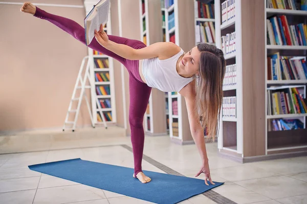 Beautiful young woman reading book and practices yoga asana Ardha Chandrasana - Half Moon pose in the library