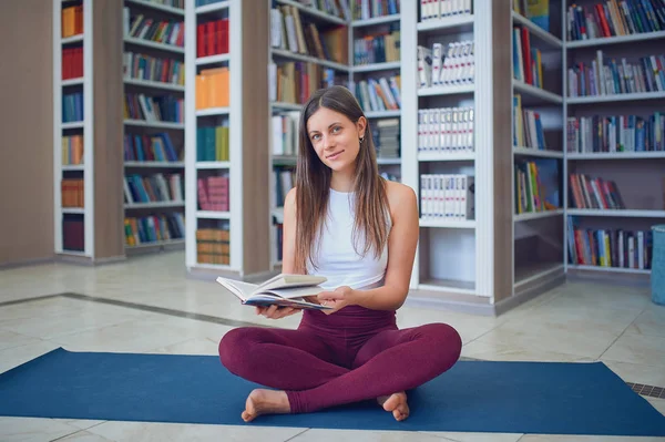 Beautiful young woman reading book and practices yoga asana Sukhasana - The Easy Sitting crosslegged Pose in the library