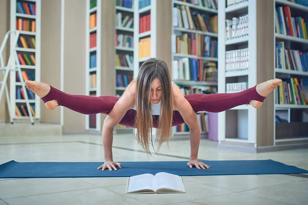 Beautiful woman reading book and practices handstand yoga asana Tittibhasana - firefly pose in the library — Stock Photo, Image