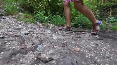 Ormanda taş bir yolda yürüyen bir kadın, yaz yürüyüşü sandaletler ve günlük kıyafetlerle, doğa patikalarını keşfeden bir kadın..