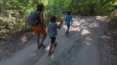 Güneşli bir günde yeşil bir ormanda aile yürüyüşü. Sırt çantalı ve çocuklu bir kadın yokuş yukarı yürüyor, doğa, seyahat ve yaz tatili aktivitelerinden keyif alıyor..