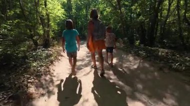 Güneşli bir günde yeşil bir ormanda aile yürüyüşü. Sırt çantalı ve çocuklu bir kadın yokuş yukarı yürüyor, doğa, seyahat ve yaz tatili aktivitelerinden keyif alıyor..