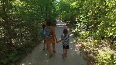 Güneşli bir günde yeşil bir ormanda aile yürüyüşü. Sırt çantalı ve çocuklu bir kadın yokuş yukarı yürüyor, doğa, seyahat ve yaz tatili aktivitelerinden keyif alıyor..