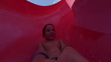 Güneşli bir günde parlak kırmızı bir su kaydırağında kayan mutlu bir çocuğun video görüntüsü. Aquapark 'ta yaz eğlencesinin tadını çıkarıyor ve sıçrıyor..