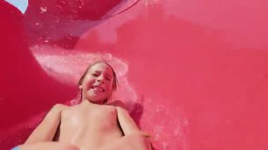 Güneşli bir günde parlak kırmızı bir su kaydırağında kayan mutlu bir çocuğun video görüntüsü. Aquapark 'ta yaz eğlencesinin tadını çıkarıyor ve sıçrıyor..