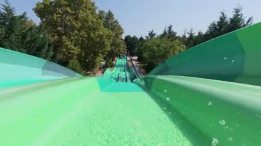 Güneşli bir günde geniş, yeşil çok şeritli bir su kaydırağında çok sayıda insan yarışıyor. Yaz su parkında eğlence ve heyecan. Sürücülerin yokuş aşağı süratle uçuşu görüntüsü.