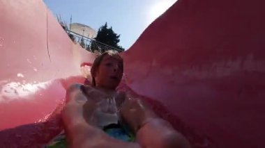 Güneşli bir günde parlak kırmızı bir su kaydırağında kayan mutlu bir çocuğun video görüntüsü. Aquapark 'ta yaz eğlencesinin tadını çıkarıyor ve sıçrıyor..