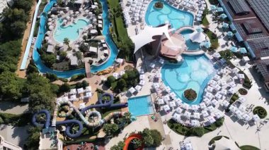 Aerial view of a luxury water park with pools, slides, lazy river, sunbeds and umbrellas on a sunny summer day, full of people relaxing and enjoying vacation activities.