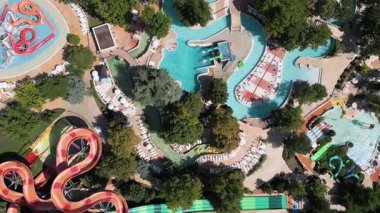 Güneşli bir günde yemyeşil ve dinlenme yerleriyle çevrili dolambaçlı slaytlar, havuzlar ve tembel nehirlerle dolu renkli bir su parkının hava aracı görüntüsü..