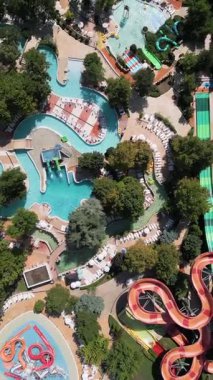 Güneşli bir günde yemyeşil ve dinlenme yerleriyle çevrili dolambaçlı slaytlar, havuzlar ve tembel nehirlerle dolu renkli bir su parkının hava aracı görüntüsü..