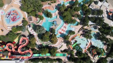 Güneşli bir günde yemyeşil ve dinlenme yerleriyle çevrili dolambaçlı slaytlar, havuzlar ve tembel nehirlerle dolu renkli bir su parkının hava aracı görüntüsü..