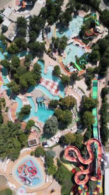 Güneşli bir günde yemyeşil ve dinlenme yerleriyle çevrili dolambaçlı slaytlar, havuzlar ve tembel nehirlerle dolu renkli bir su parkının hava aracı görüntüsü..