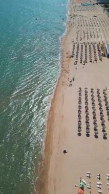 Kıyı boyunca sıralı şemsiyeler ve güneş yatakları olan kalabalık bir sahil manzarası ve güneşli bir yaz gününde açık turkuaz denizde yüzen insanlar..