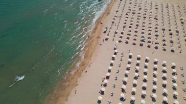 Kıyı boyunca sıralı şemsiyeler ve güneş yatakları olan kalabalık bir sahil manzarası ve güneşli bir yaz gününde açık turkuaz denizde yüzen insanlar..