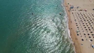 Kıyı boyunca sıralı şemsiyeler ve güneş yatakları olan kalabalık bir sahil manzarası ve güneşli bir yaz gününde açık turkuaz denizde yüzen insanlar..